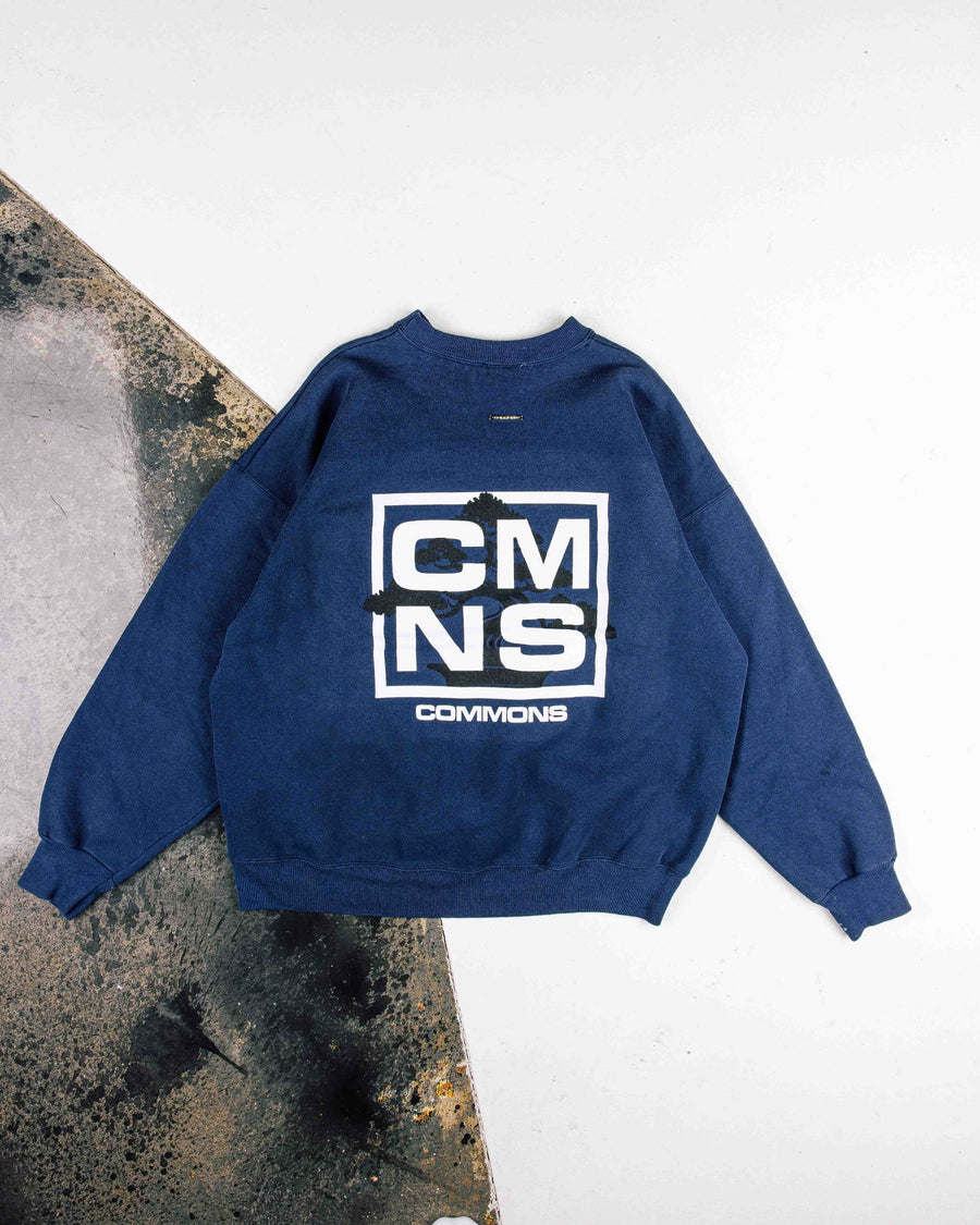 Navy crewneck sweatshirt with white CMNS Commons lettering and a black tree graphic, laid flat on white and worn concrete.