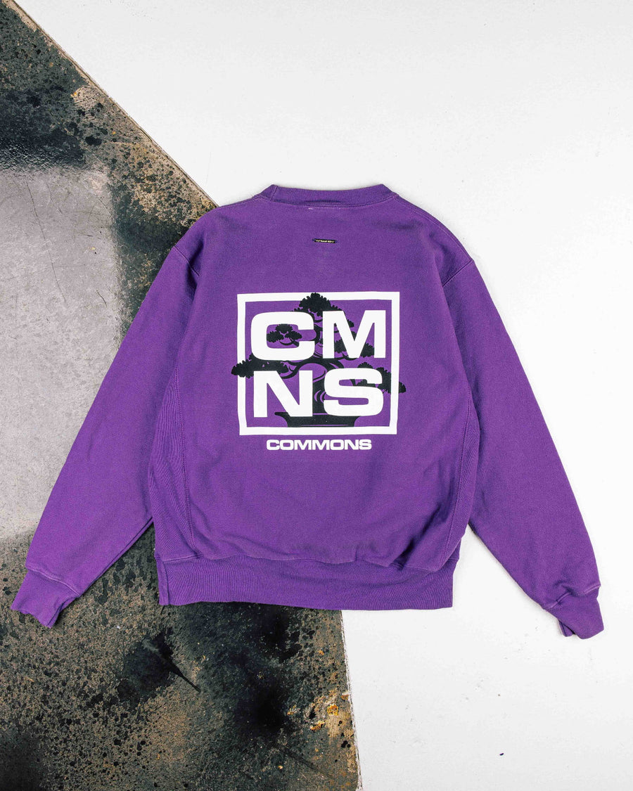 Purple crewneck sweatshirt laid flat, showing a white CMNS Commons logo and black bonsai graphic on the back.