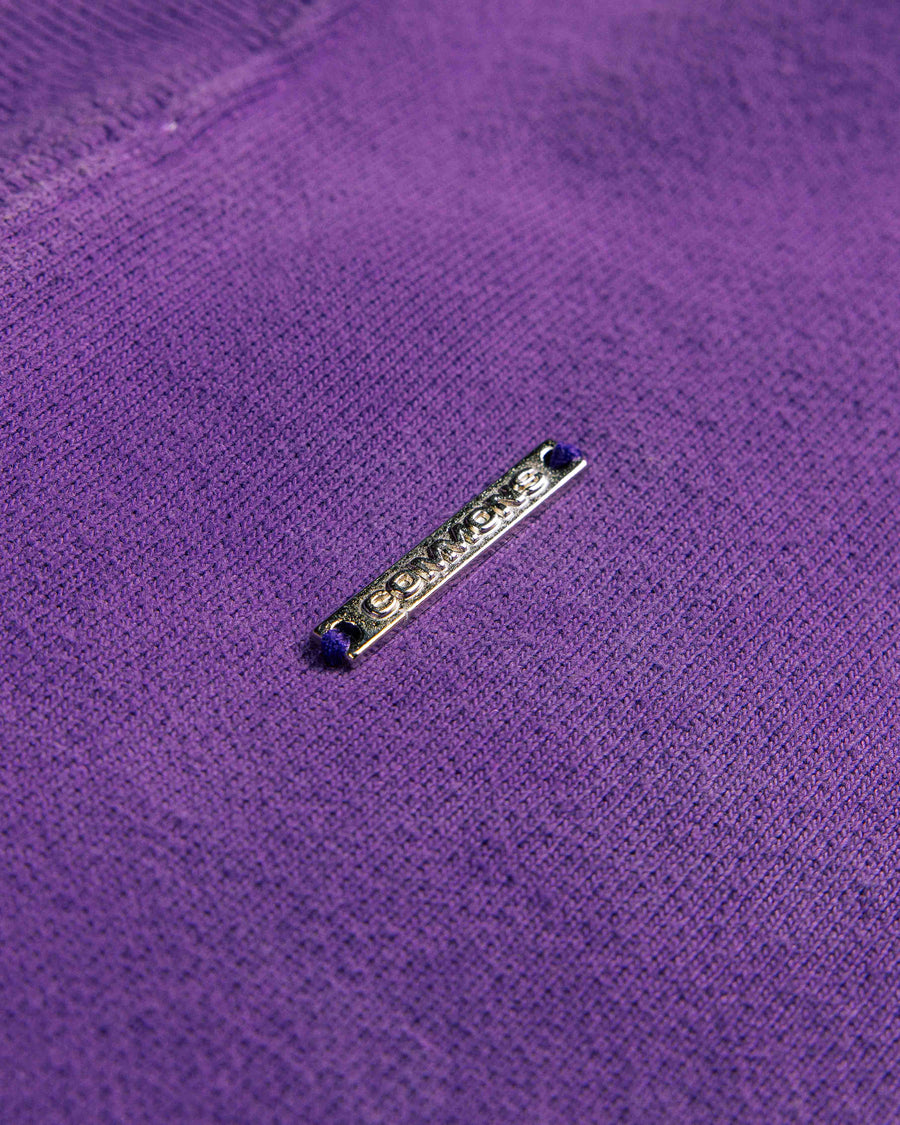 Close-up of purple knit fabric with a slim silver-tone metal logo tag stitched in place.
