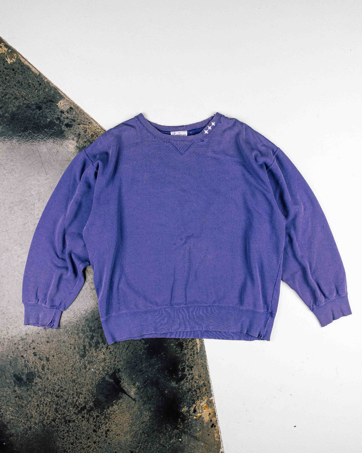 Purple crewneck sweatshirt with white stars at the collar and ribbed cuffs, laid flat on a white and weathered surface.