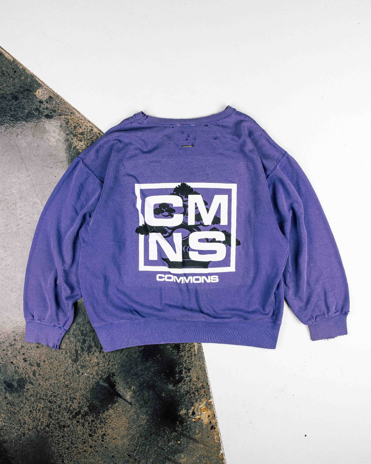 Purple crewneck sweatshirt with distressed edges, white CMNS lettering and a black bonsai graphic, laid flat.