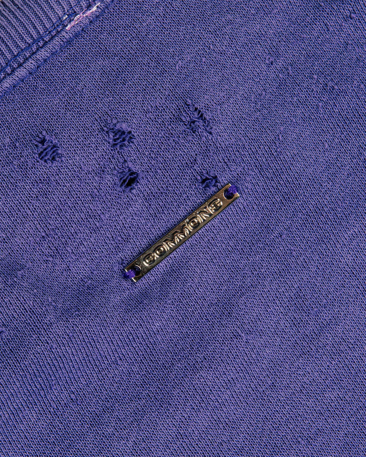 Close-up of a purple knit top with distressed holes, ribbed neckline, and a gold-tone COMMONS metal tag.