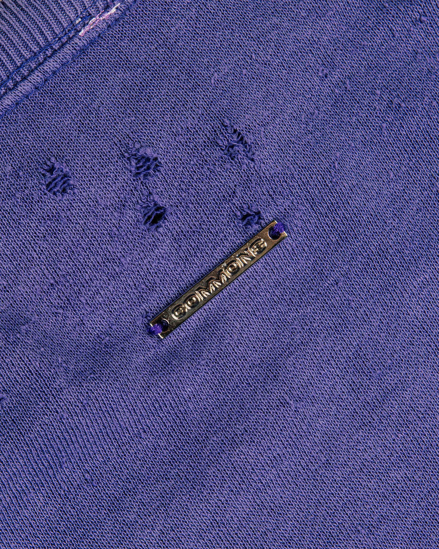 Close-up of a purple knit top with distressed holes, ribbed neckline, and a gold-tone COMMONS metal tag.