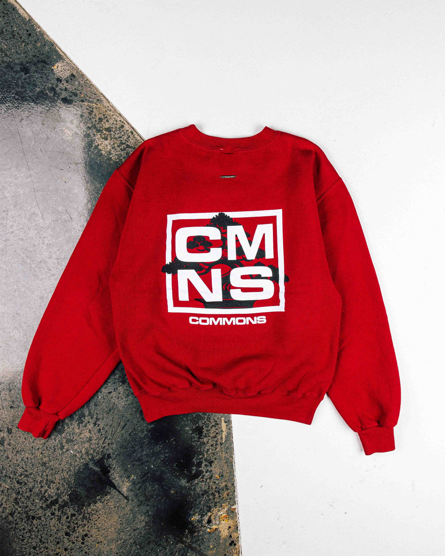 Red crewneck sweatshirt with white CMNS lettering and a black tree graphic, laid flat on a white and weathered gray surface.