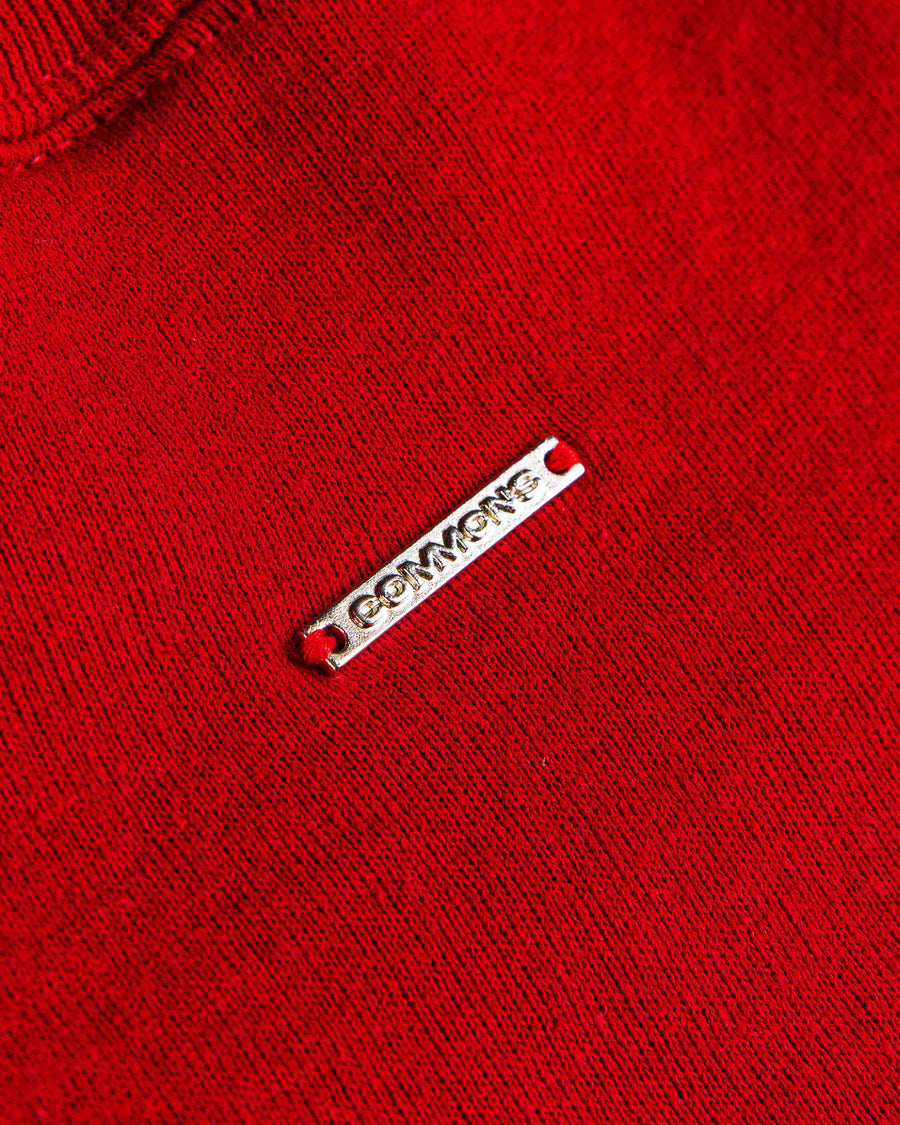 Close-up of a red knit sweater with a ribbed neckline and a small silver-tone metal logo tag.
