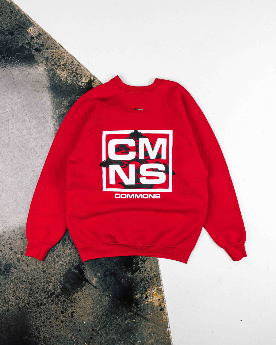Red crewneck sweatshirt with white CMNS lettering and a black tree graphic, laid flat on a white and weathered gray surface.