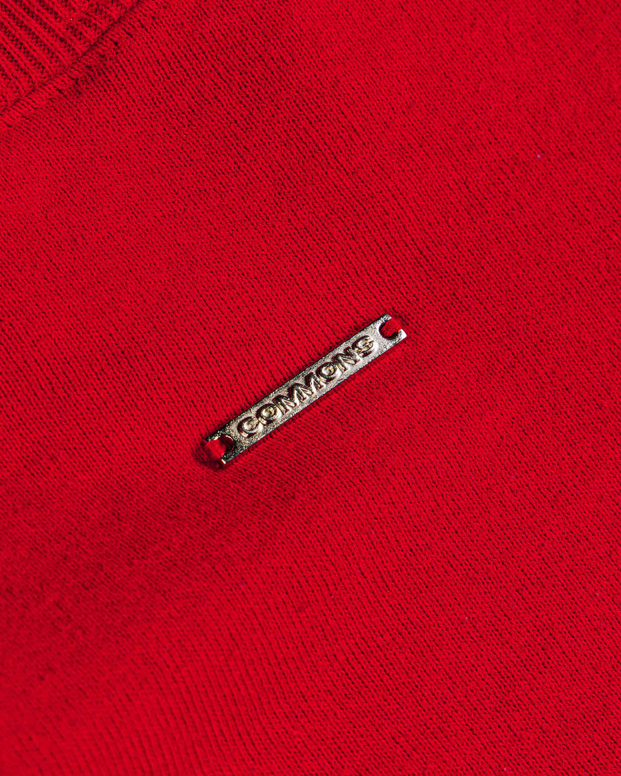 Close-up of red knit fabric with ribbed trim and a silver-tone metal logo tag stitched in place.
