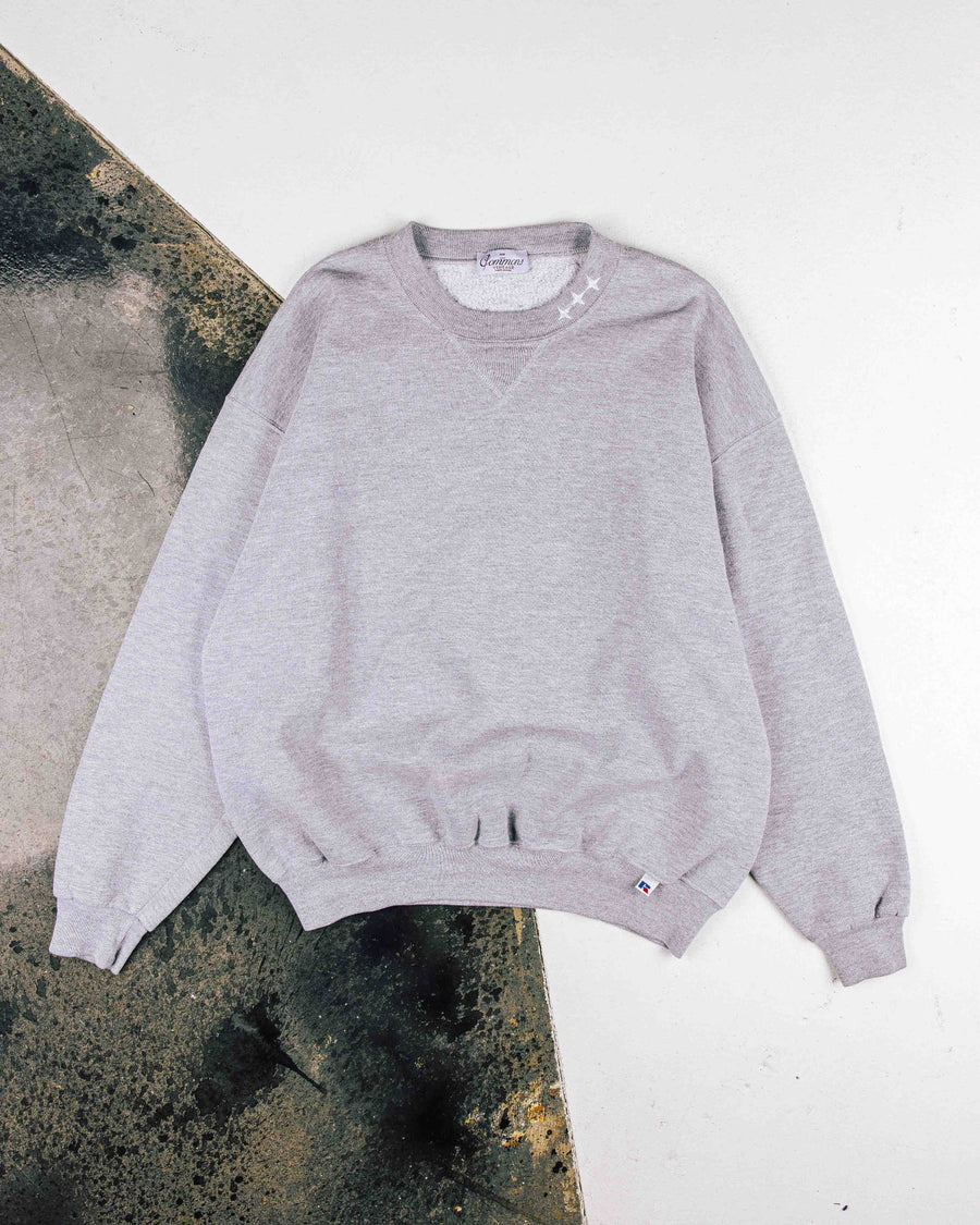 Light gray crewneck sweatshirt with ribbed cuffs, white collar stitching, and a small hem logo, laid flat on concrete.