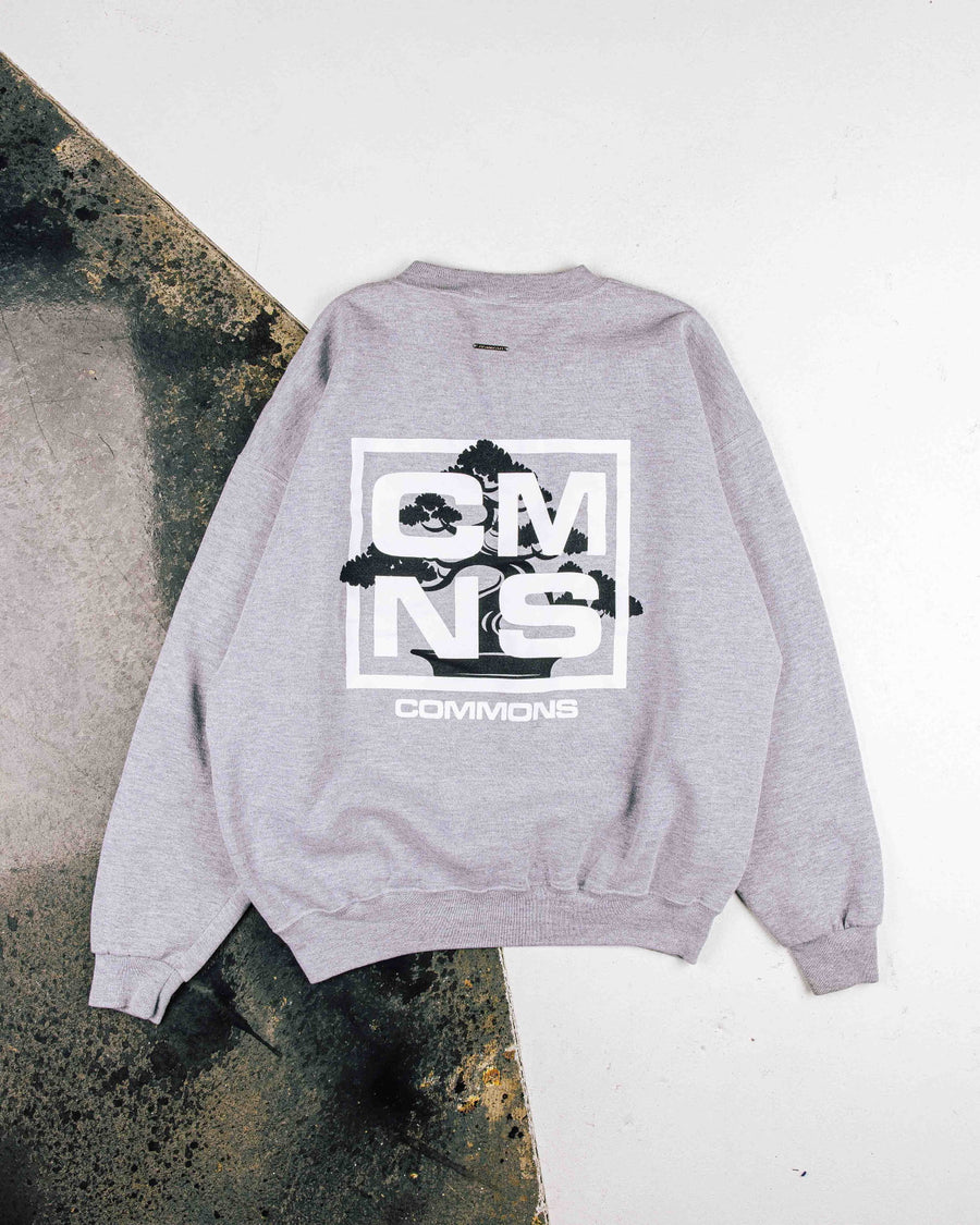 Gray crewneck sweatshirt with white CMNS lettering and a black bonsai back graphic, laid flat on a two-tone surface.