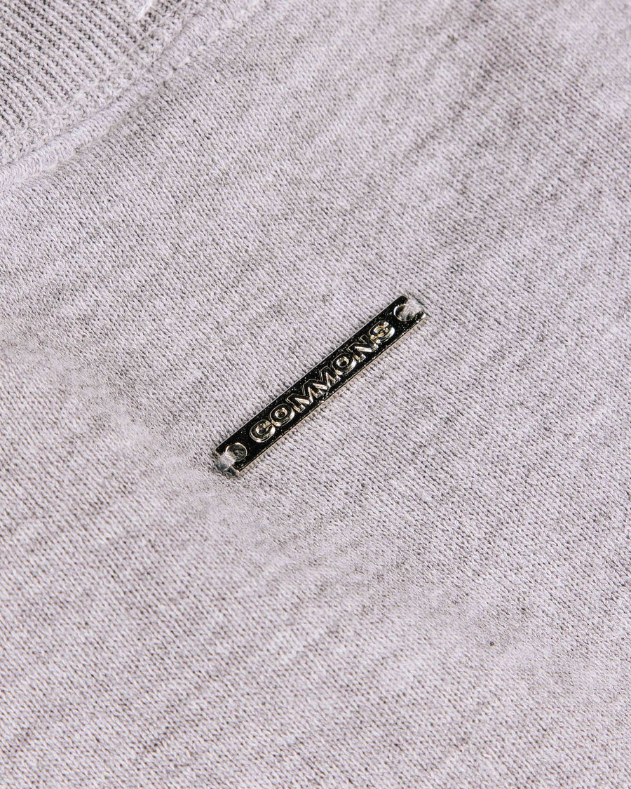 Close-up of a light gray knit garment with ribbed trim and a rectangular metal COMMONS logo tag.