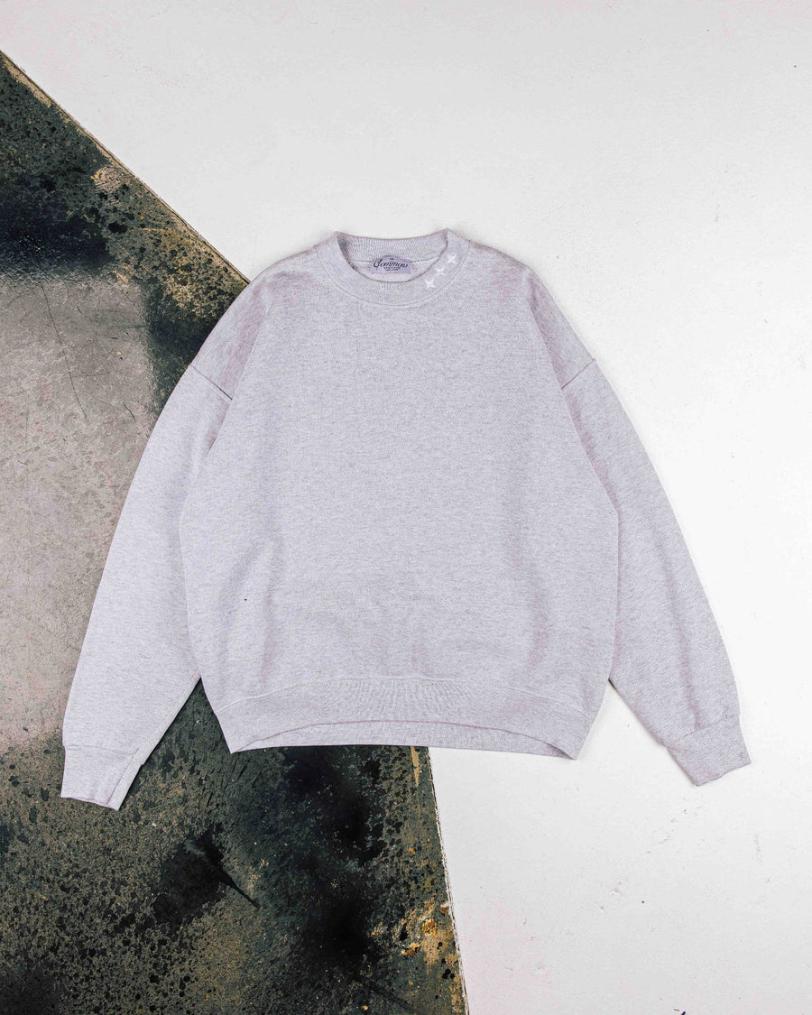 Light gray crewneck sweatshirt with ribbed cuffs and hem and white stars near the collar, laid flat on a two-tone surface.