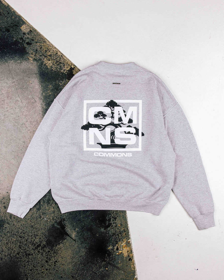 Gray sweatshirt laid flat, showing a black bonsai graphic and white CMNS lettering on the back, with ribbed cuffs and hem.