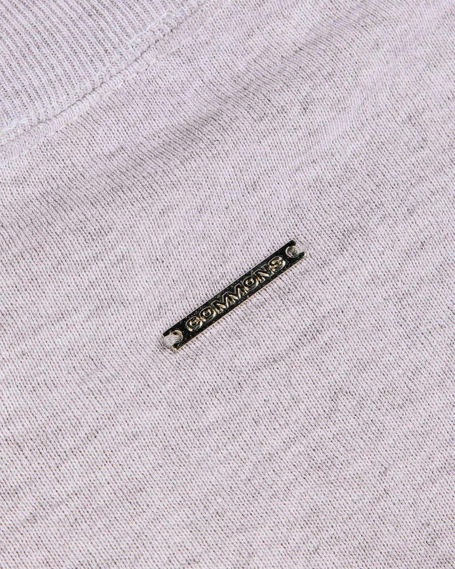 Close-up of a light gray knit garment with ribbed trim and a rectangular metal COMMONS logo tag.