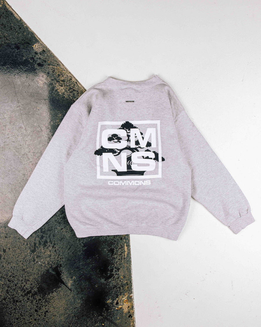 Light gray crewneck sweatshirt with a black bonsai graphic and white COMMONS lettering, laid flat on a two-tone surface.