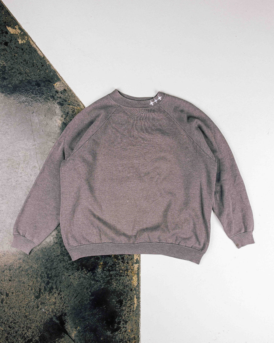 Gray raglan sweatshirt with white stars at the ribbed crewneck, laid flat on a white and distressed dark surface.
