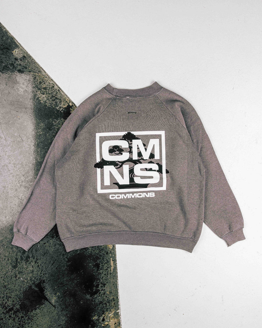 Gray sweatshirt laid flat on a two-tone floor, with a black bonsai graphic and white CMNS and COMMONS lettering on the back.