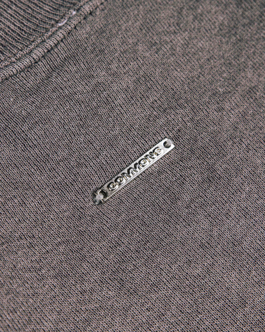 Close-up of a gray knit top with a ribbed neckline and a silver-tone metal logo tag stitched onto the fabric.