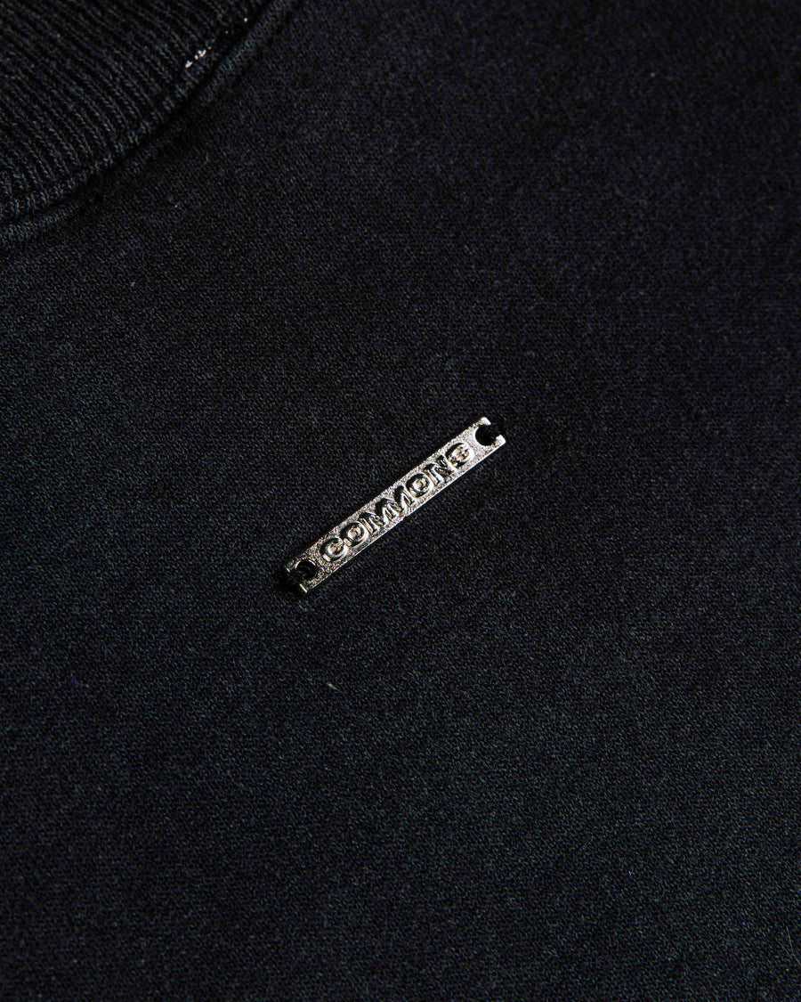 Close-up of a black knit top with a ribbed neckline and a silver-tone COMMONS logo plaque.