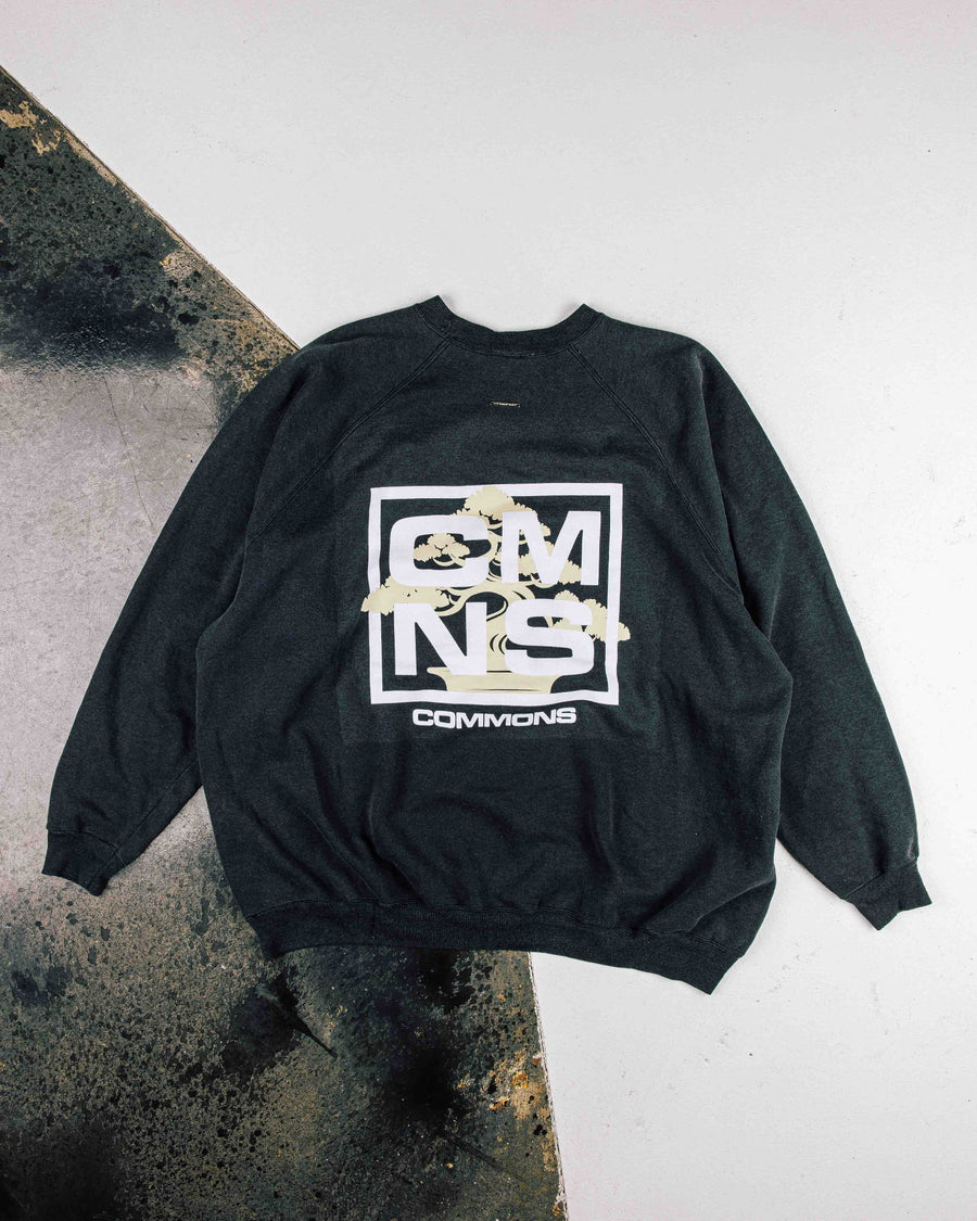 Charcoal crewneck sweatshirt with white CMNS lettering and a pale yellow bonsai graphic, laid flat on a two-tone floor.