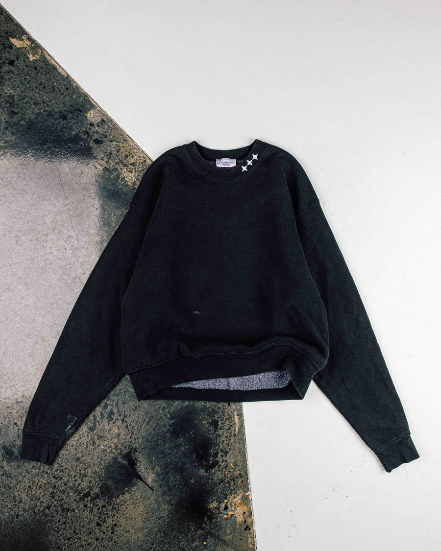 Black crewneck sweatshirt with three white stars at the collar, laid flat on a white and weathered concrete surface.
