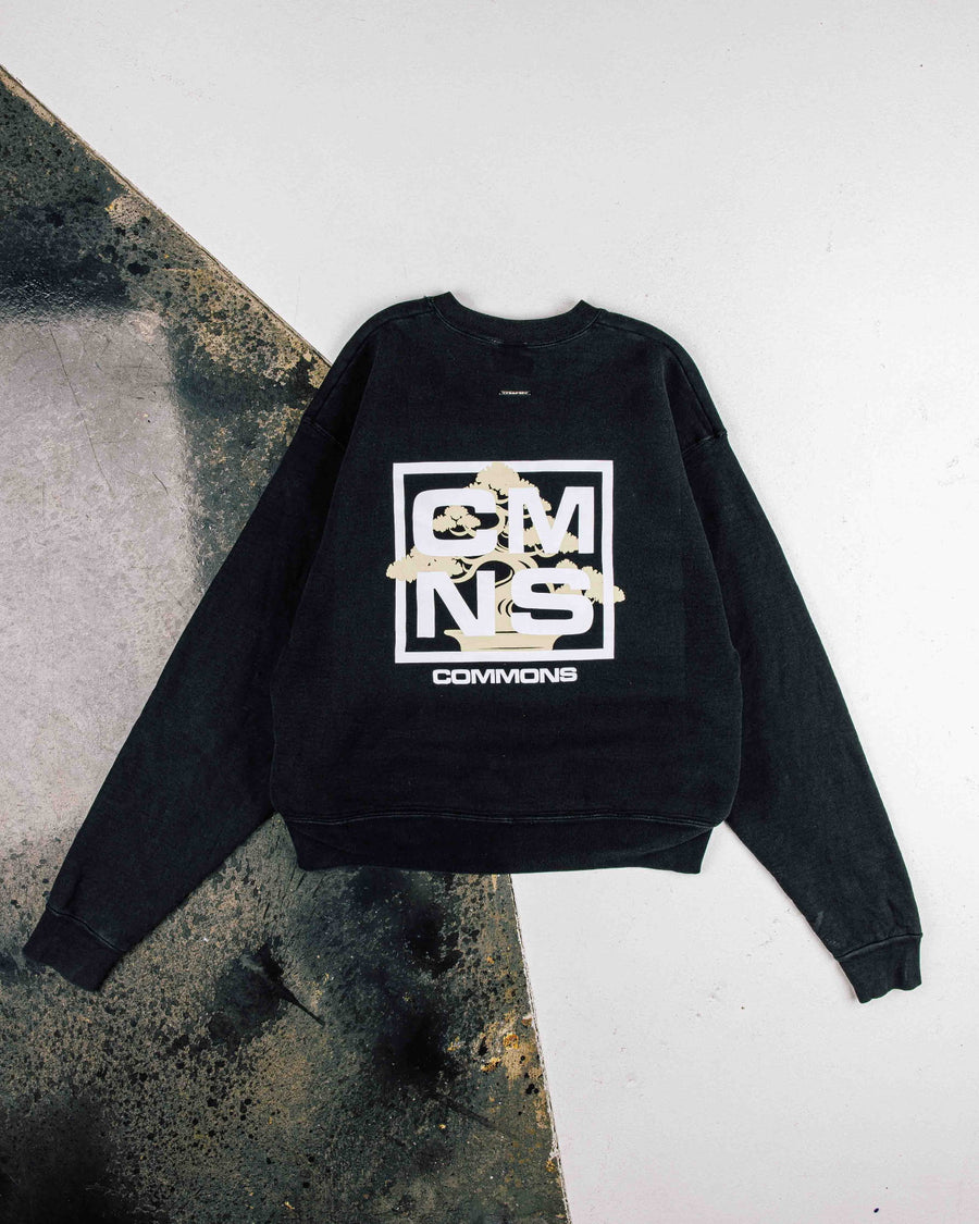 Black crewneck sweatshirt with white COMMONS lettering and a beige bonsai graphic, laid flat on a two-tone surface.