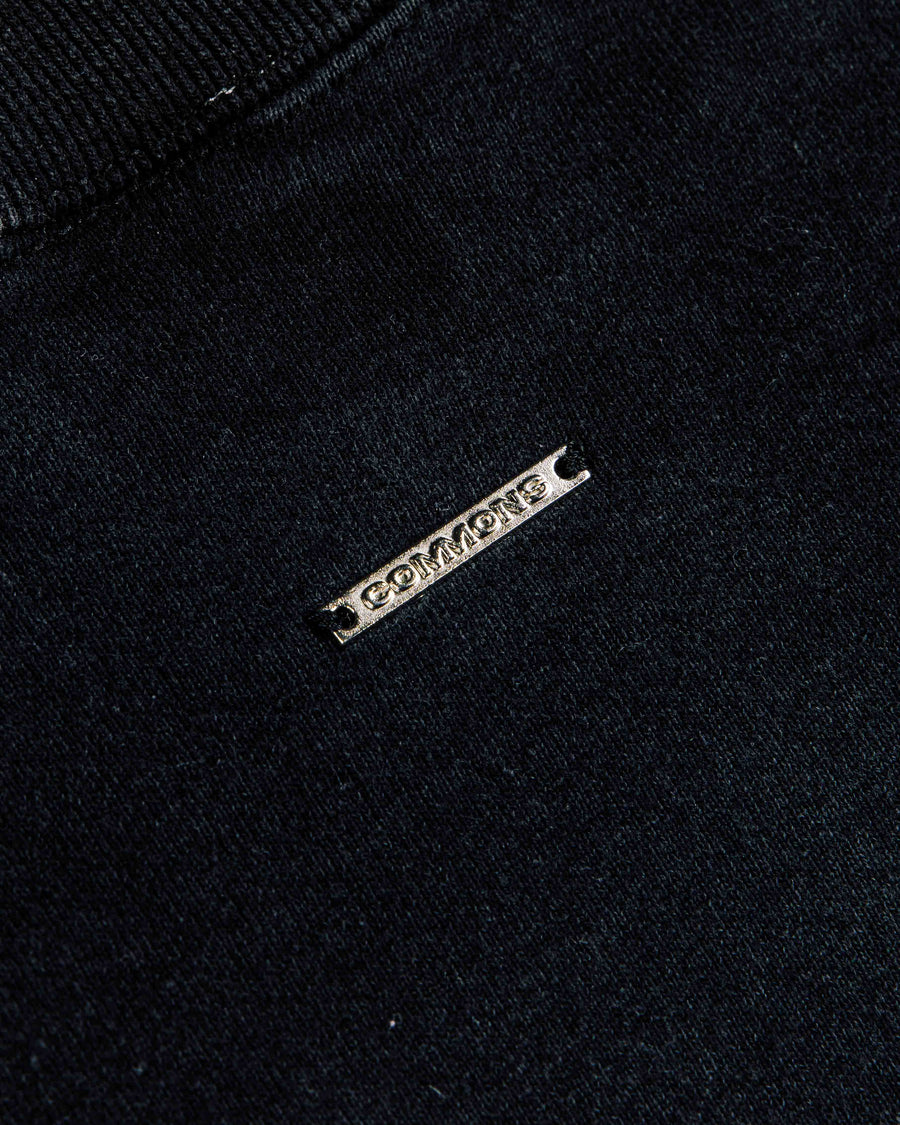 Close-up of black knit clothing with ribbed trim and a silver-tone metal COMMONS logo tag.