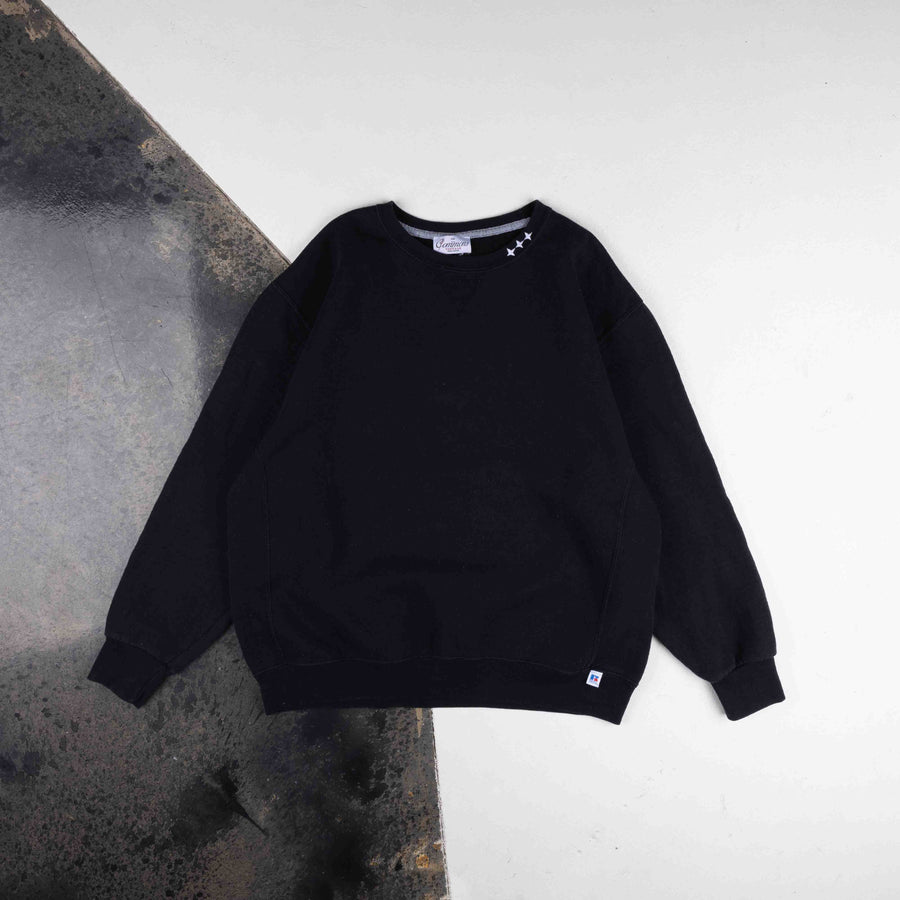 Black crewneck sweatshirt with white stars at the collar and ribbed cuffs, laid flat on a white and gray surface.