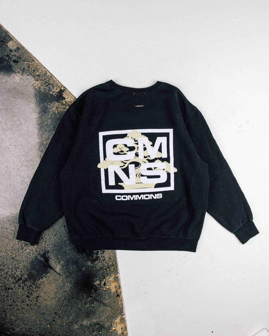 Black crewneck sweatshirt with white lettering and a pale yellow bonsai graphic, laid flat on a white and gray surface.