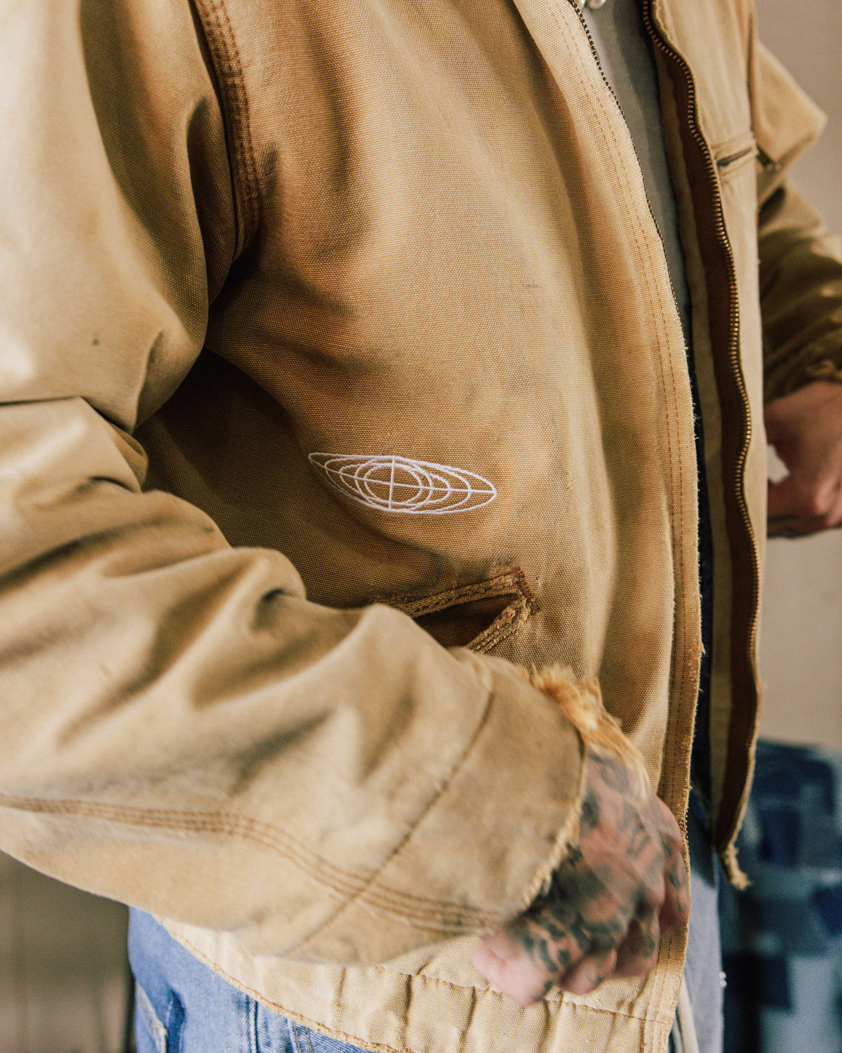 Tan canvas zip-front jacket with white embroidered emblem, frayed pocket and cuffs, worn with blue jeans.
