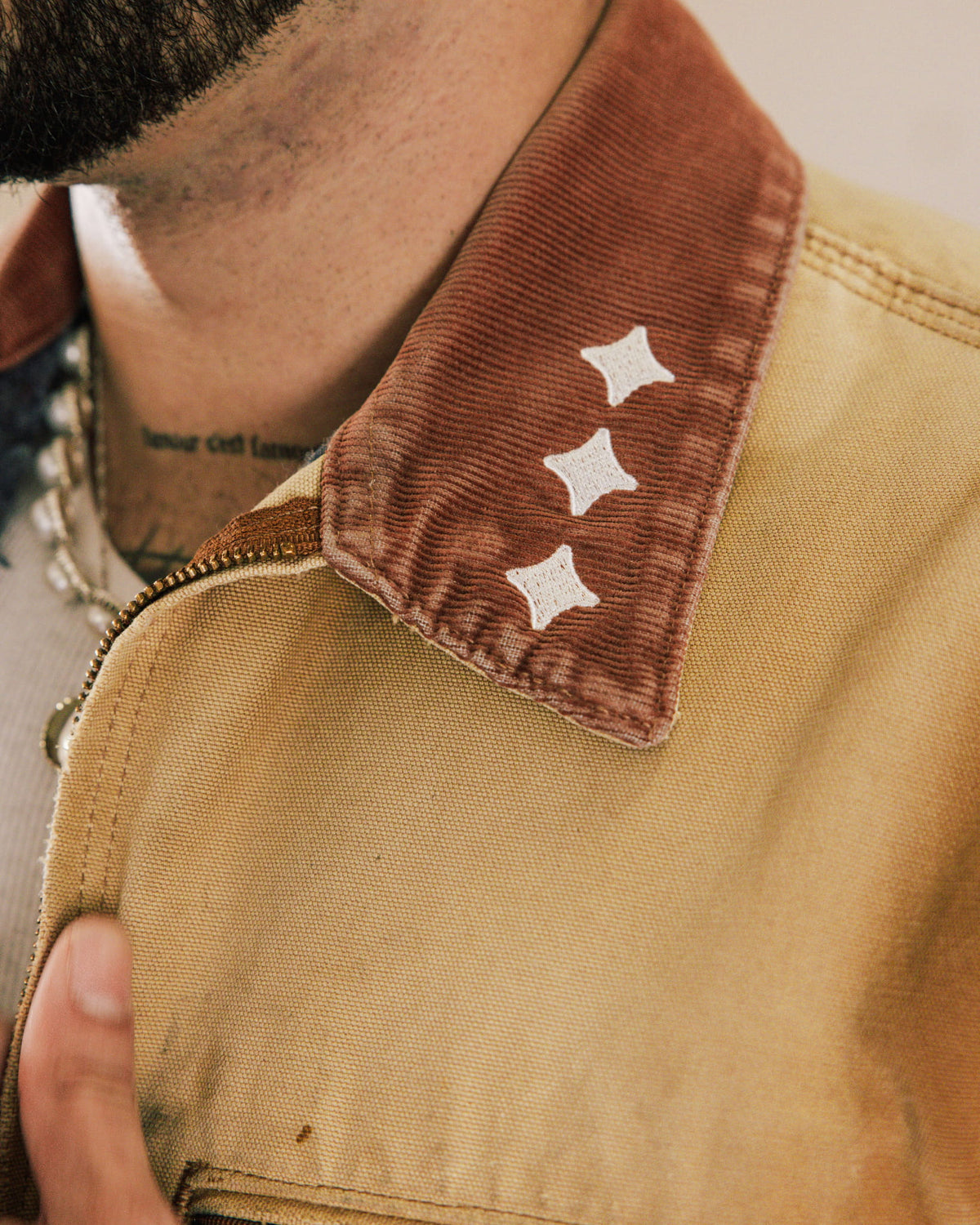 Close-up of a tan canvas jacket with a brown corduroy collar, three white embroidered stars, and a metal zipper.