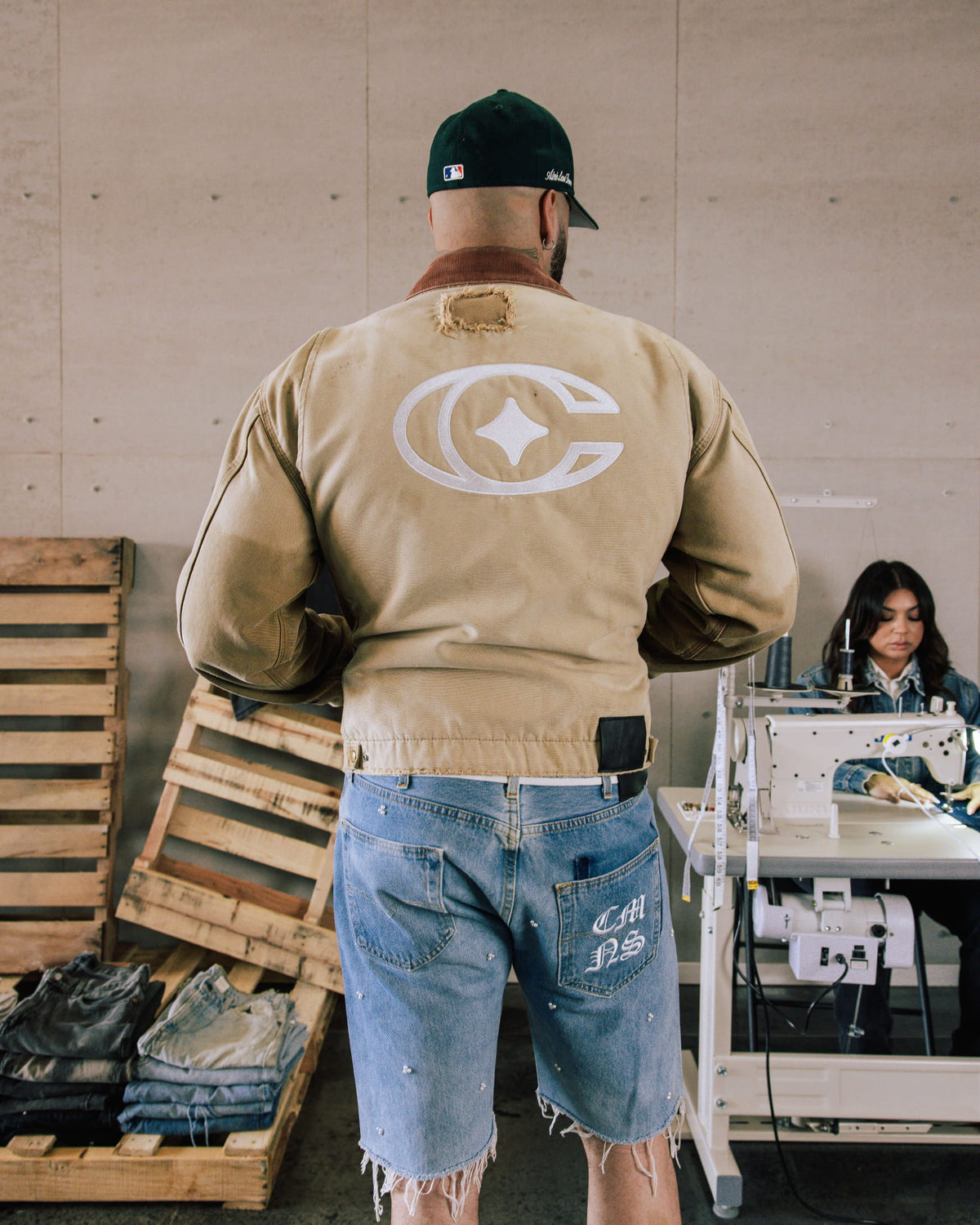 Man wearing a tan jacket with a white back logo and brown collar, plus frayed blue denim shorts in a sewing workshop.