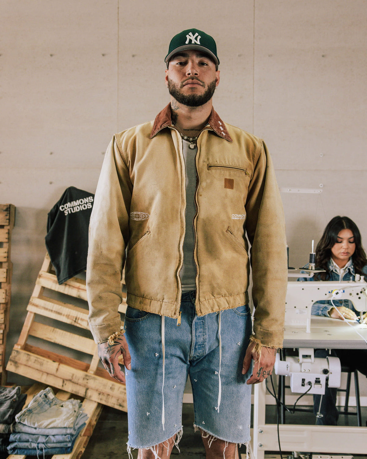 Man wearing a tan zip jacket with brown collar, frayed denim shorts and a green cap in a sewing workshop.