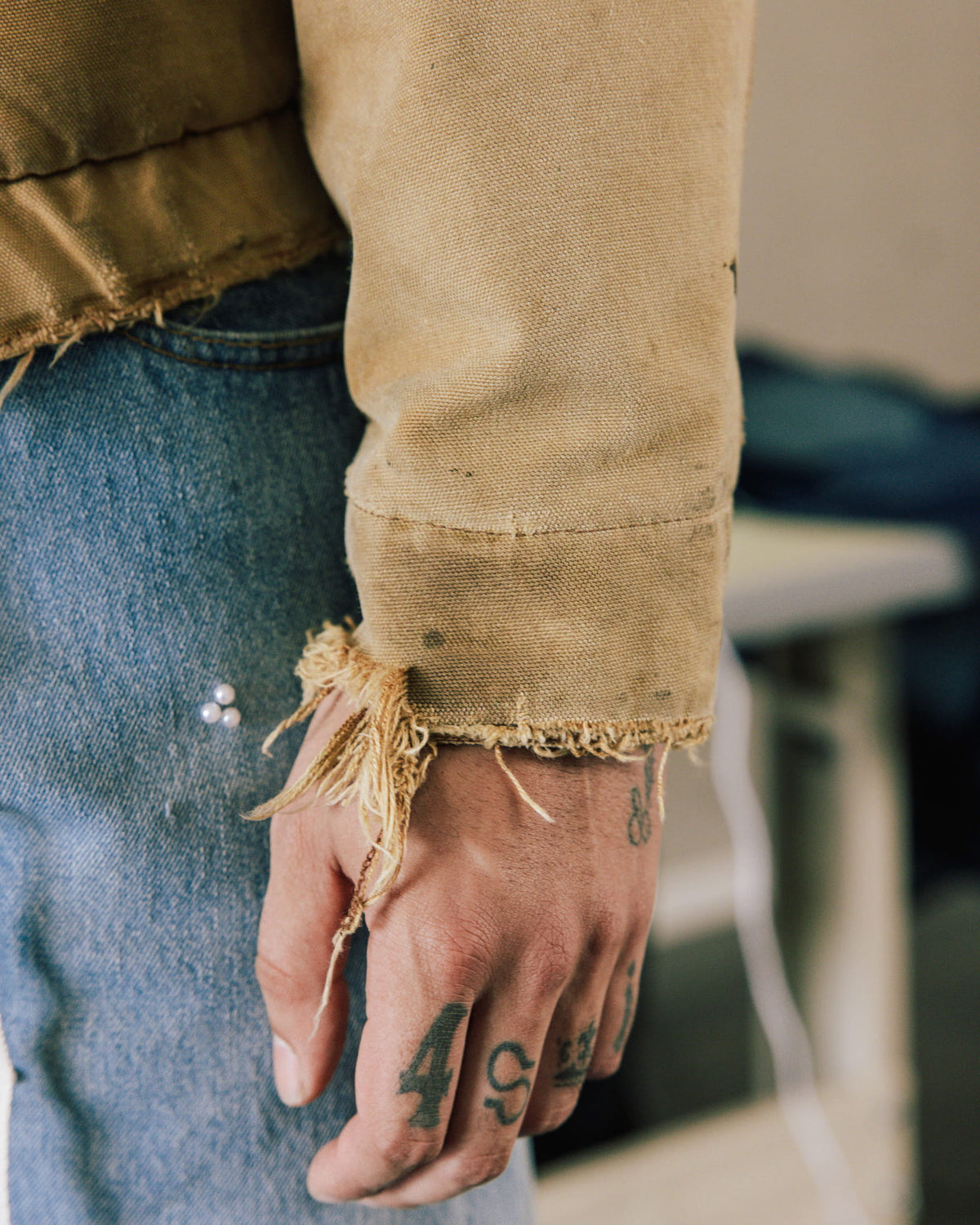 Close-up of a tan canvas jacket with a frayed cuff, worn with blue jeans beside a tattooed hand.