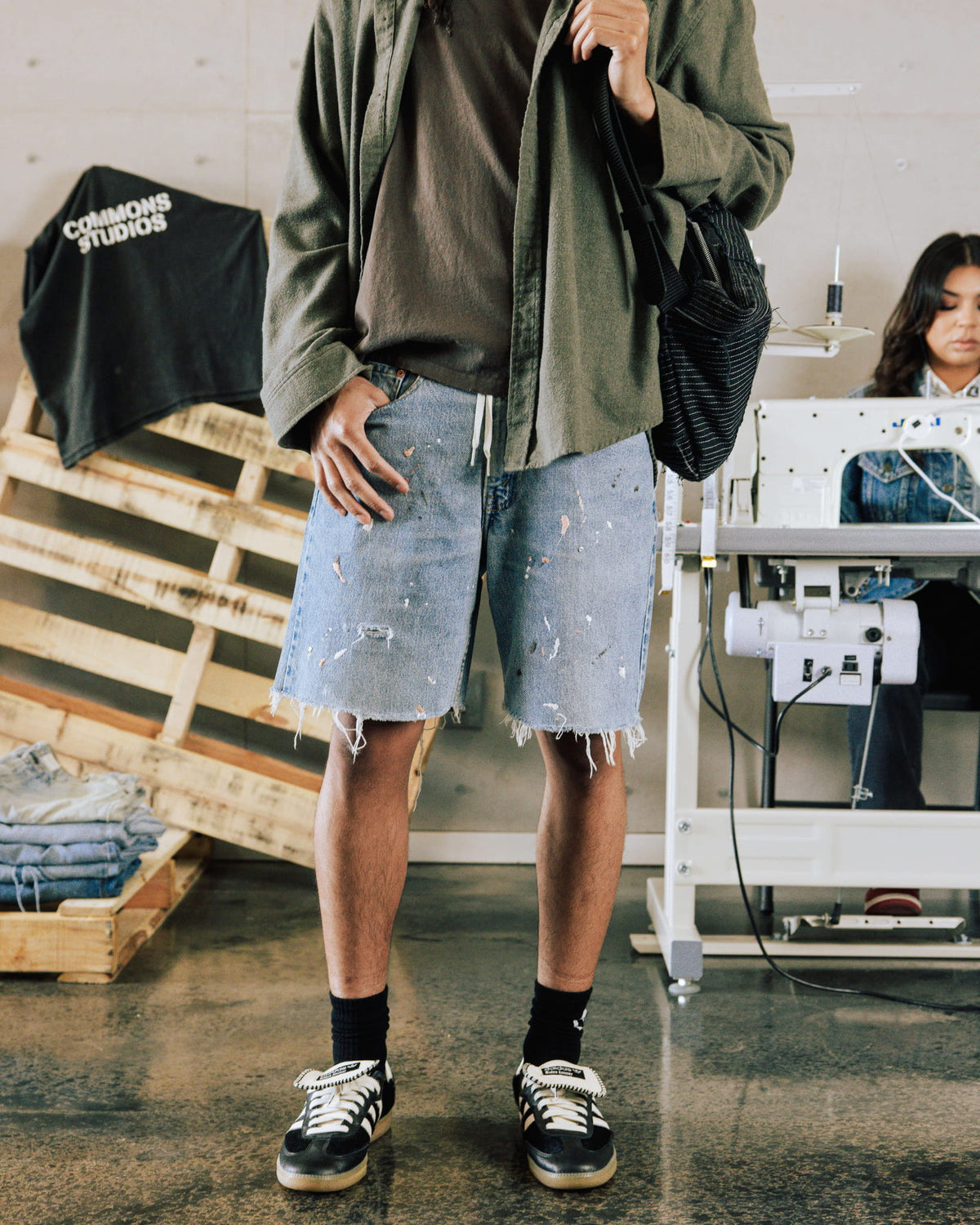 Model wears light blue paint-splattered denim shorts with frayed hems and an olive overshirt in a sewing studio.