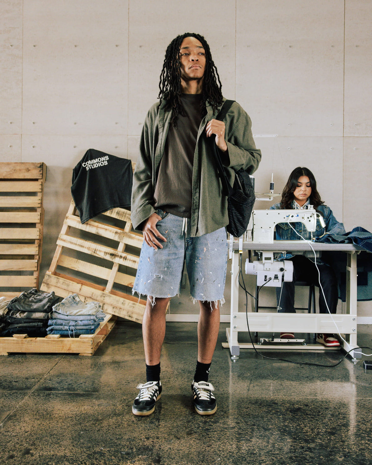 Model wears an olive overshirt, dark tee and distressed light blue denim shorts with a black shoulder bag in a sewing studio.