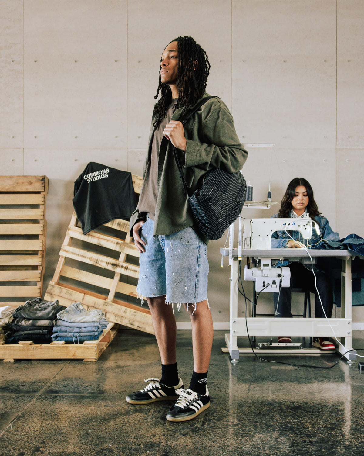 Model wears an olive overshirt, frayed denim shorts and black sneakers, carrying a black striped bag in a sewing studio.