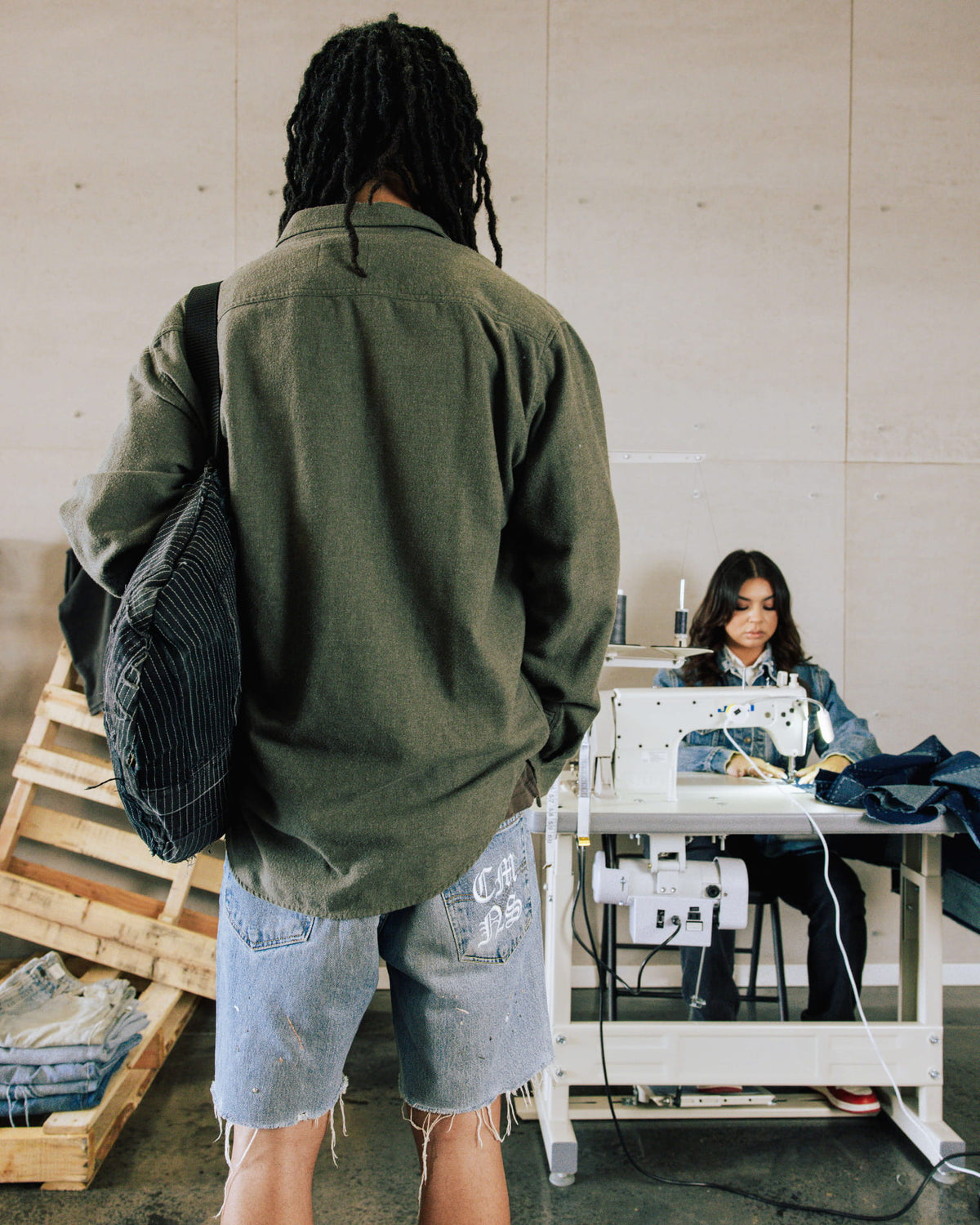 Person wearing an olive shirt, frayed denim shorts and a black striped shoulder bag in a sewing workshop.