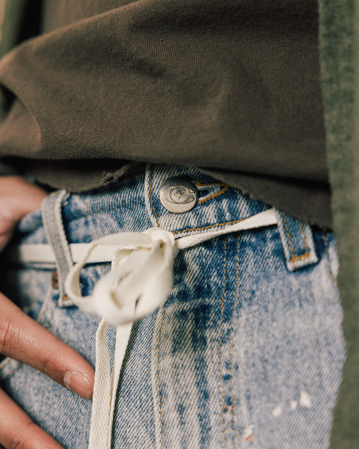 Close-up of light blue denim jeans with a metal button and cream cord tied through belt loops beneath a dark olive shirt.