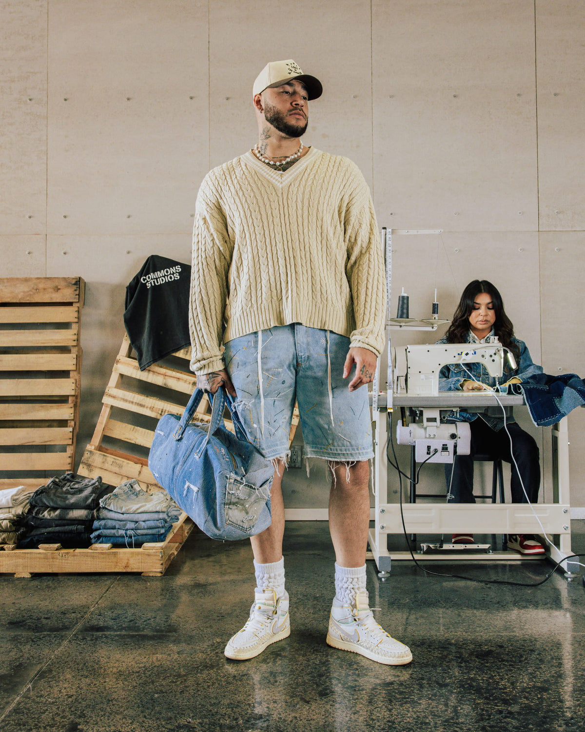 Man wearing a cream cable-knit sweater and frayed denim shorts, holding a denim duffel bag in a sewing workshop.