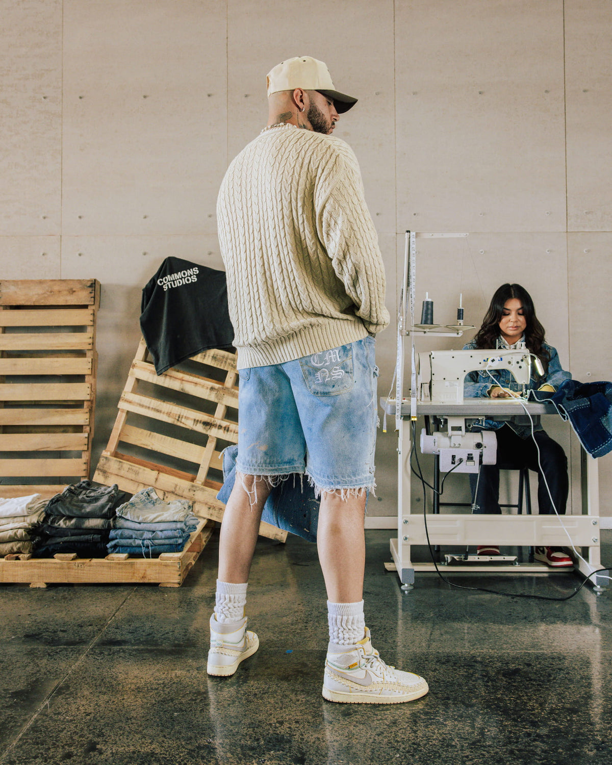 Man wearing a cream cable-knit sweater, frayed light blue denim shorts and white sneakers in a sewing workshop.
