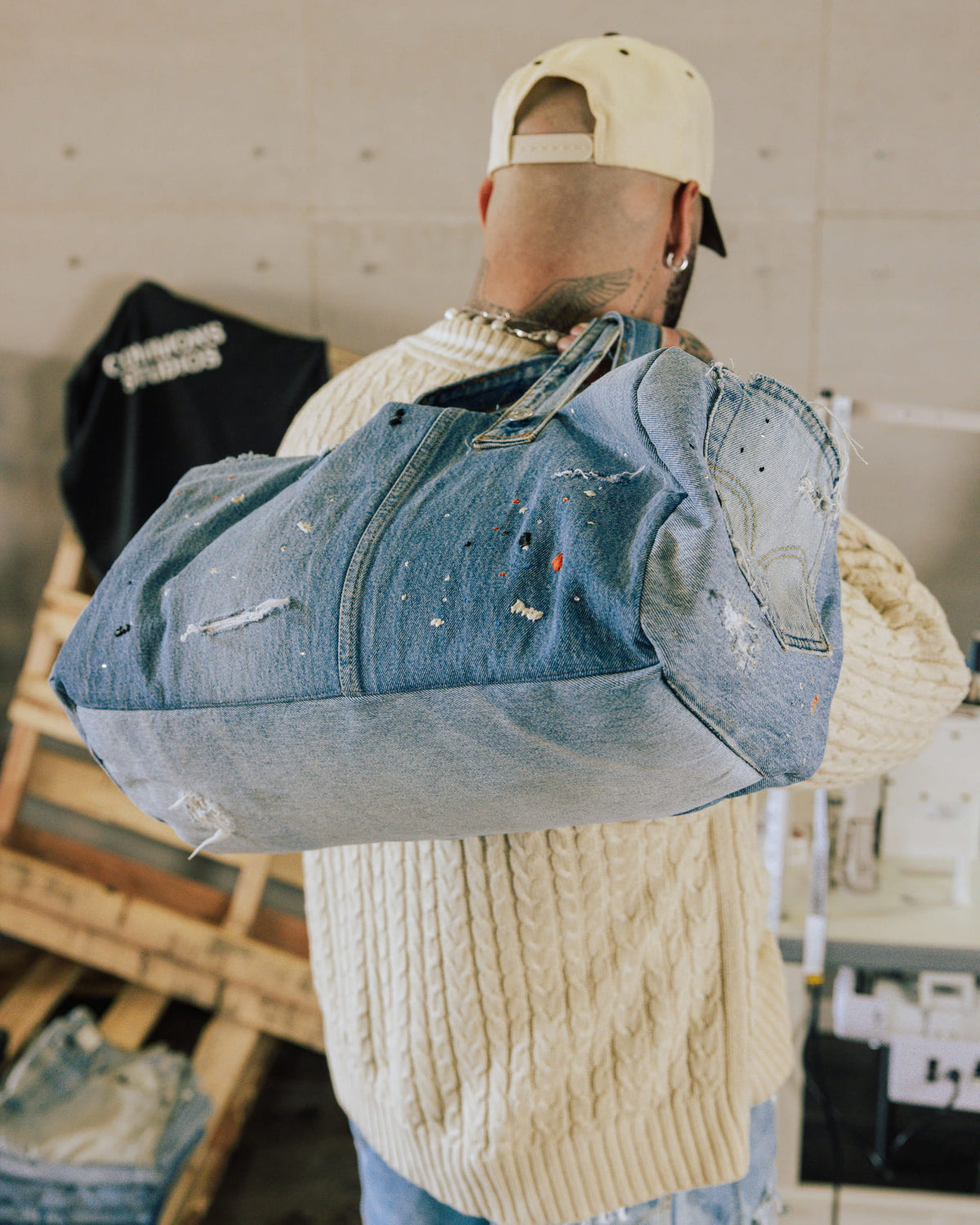 Person in a cream sweater carrying a large blue patchwork denim duffel bag with distressed details and paint splatters.