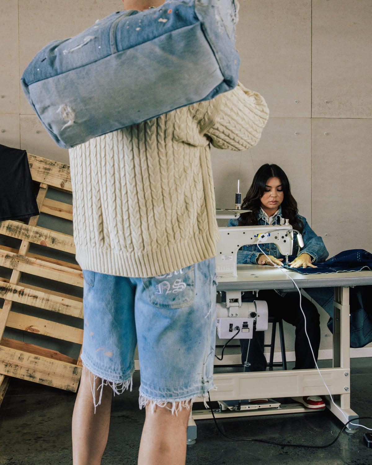 Person in a cream cable-knit sweater and frayed denim shorts carries a distressed blue denim bag in a sewing workshop.