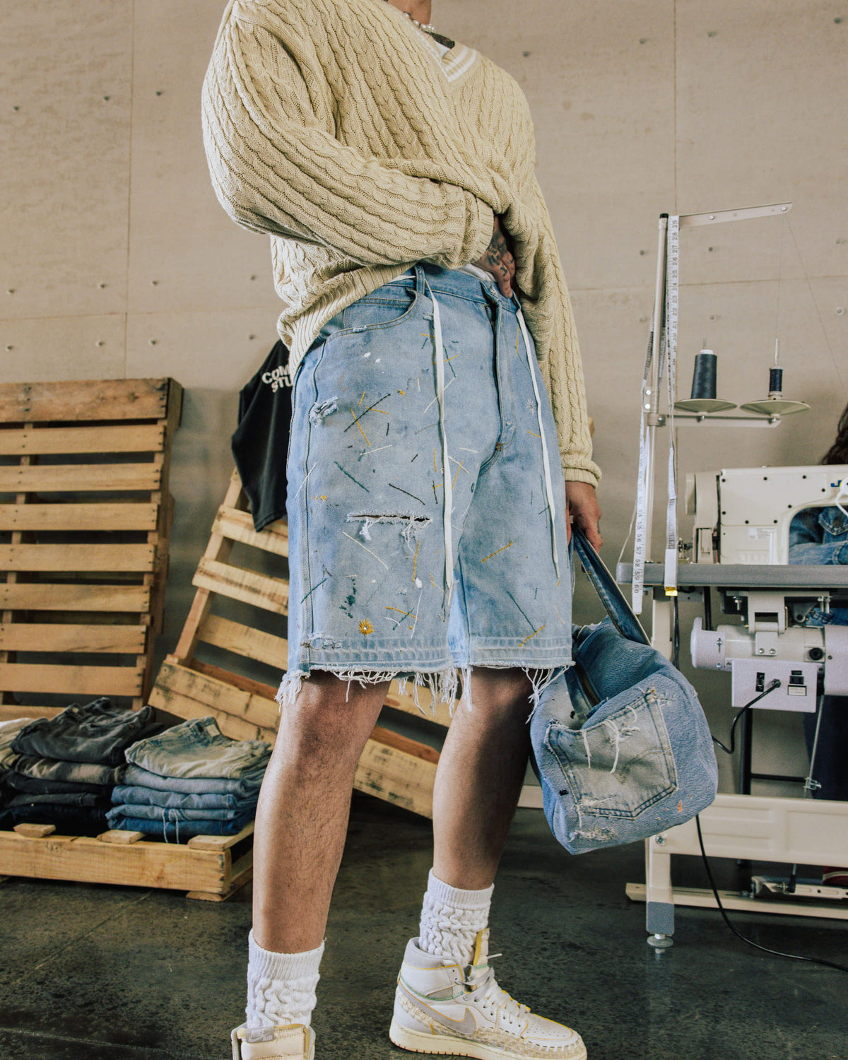 Person wearing a cream cable-knit sweater and distressed blue denim shorts, holding a denim bag in a sewing workshop.
