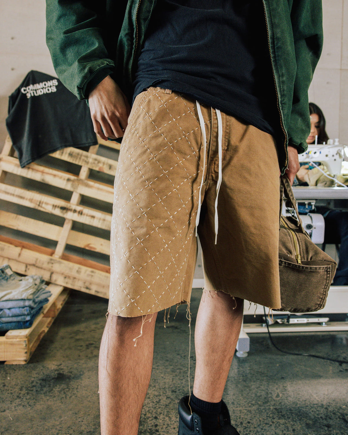 Person wearing tan drawstring shorts with white diamond stitching and frayed hems in a sewing workshop.