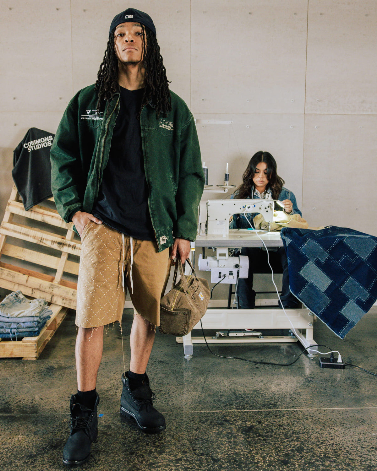 Model wears a dark green zip jacket, tan stitched shorts and black boots, holding a brown bag in a sewing workshop.