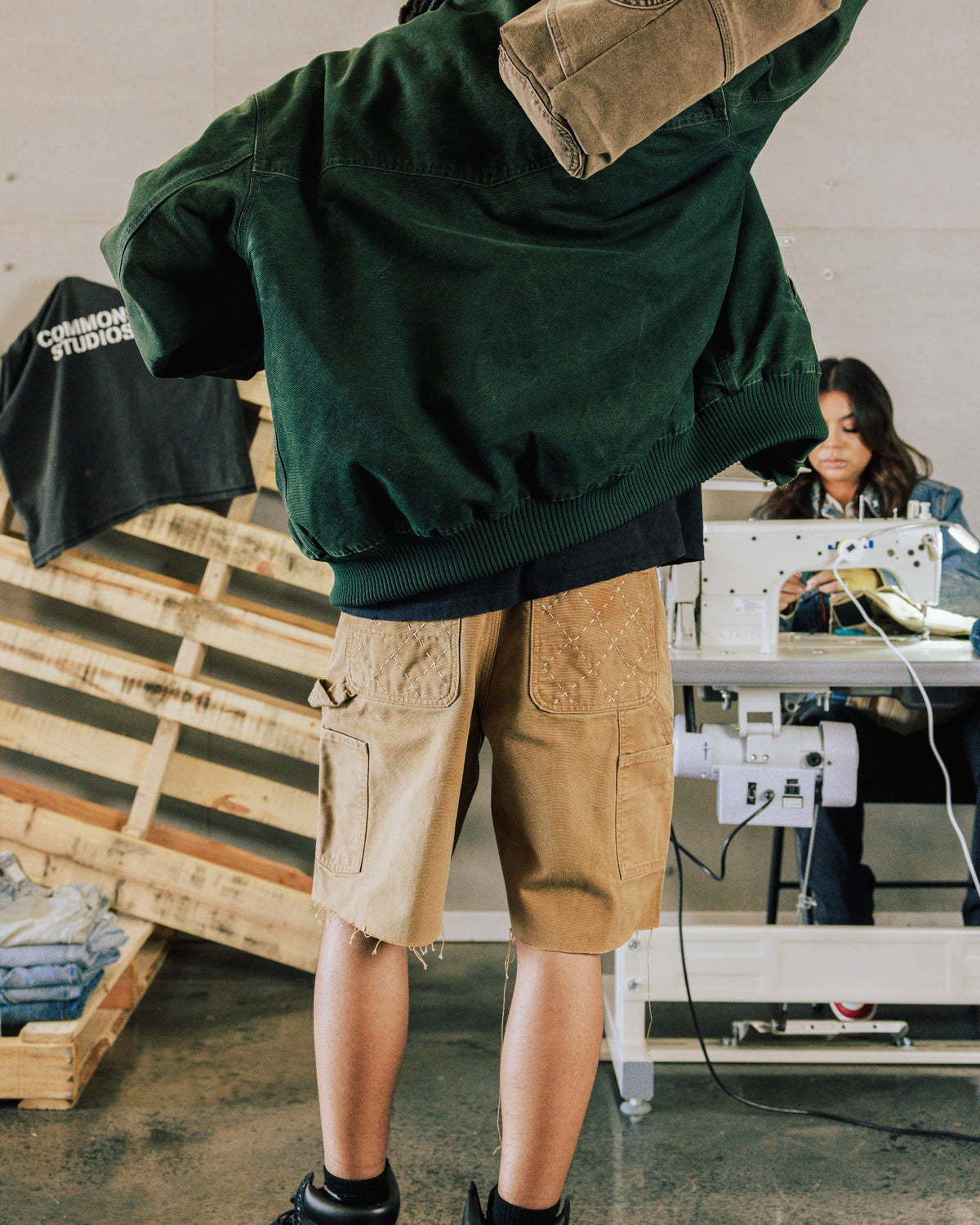 Person wearing a dark green jacket and tan cargo shorts with stitched back pockets and raw hems in a sewing workshop.