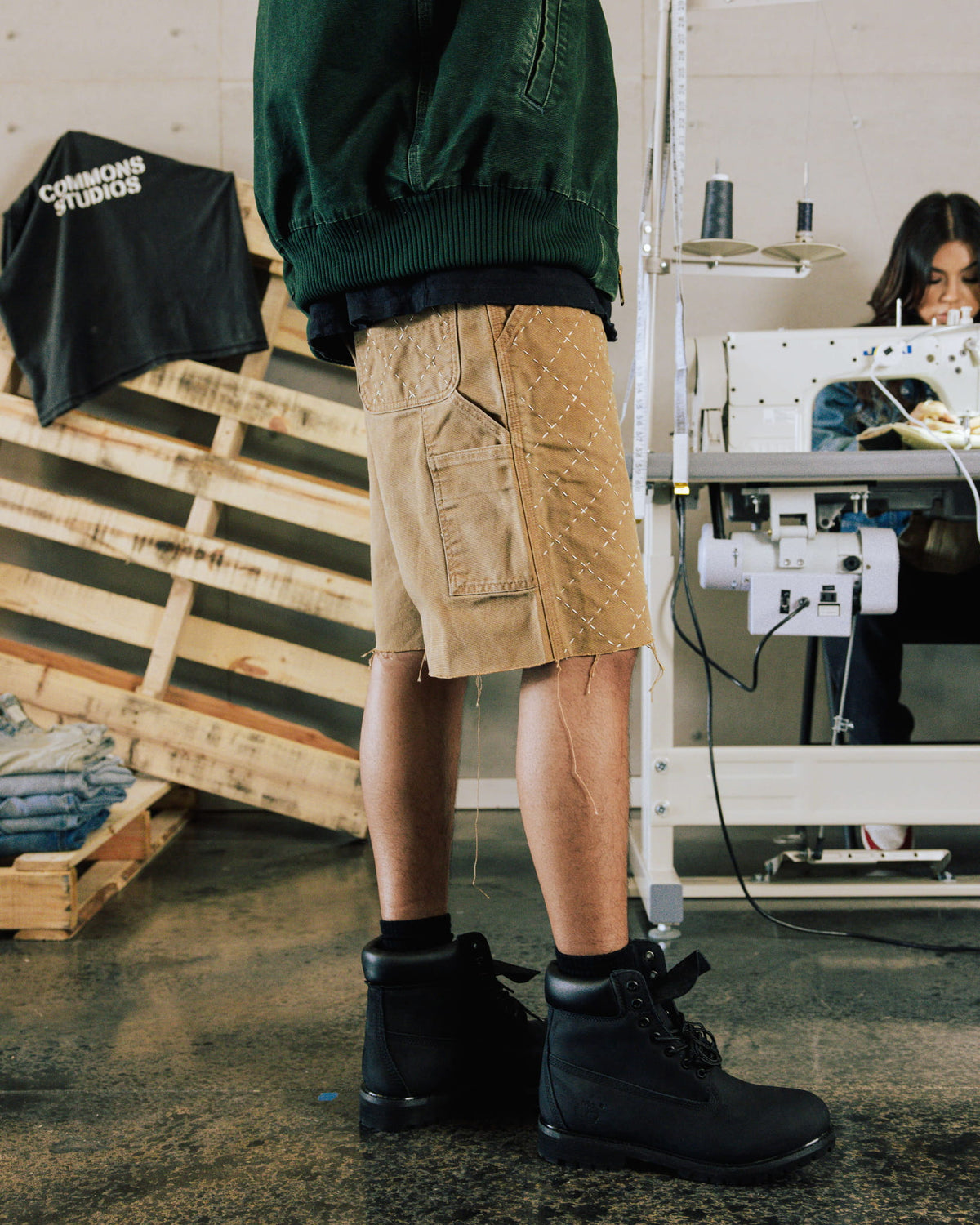 Person wearing tan shorts with white stitching, raw hems and utility pockets, a green jacket and black boots in a workshop.