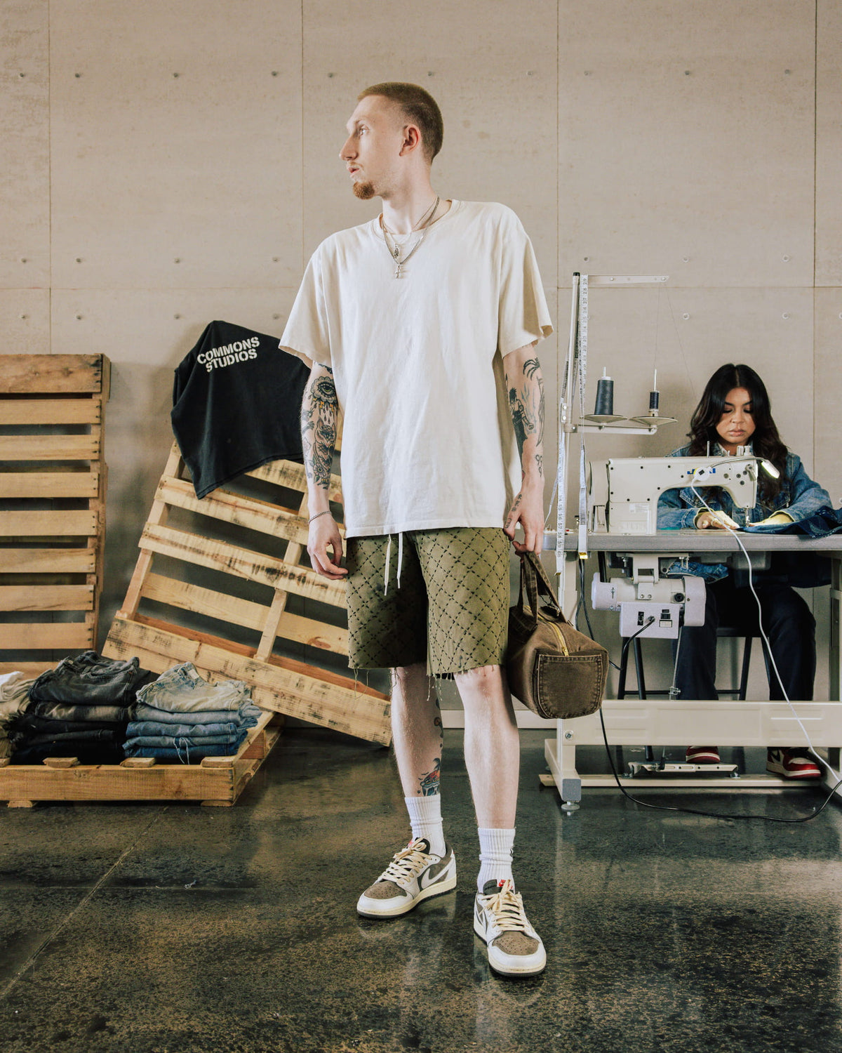 Man wearing an oversized cream T-shirt, olive patterned shorts and sneakers, holding a brown bag in a sewing studio.