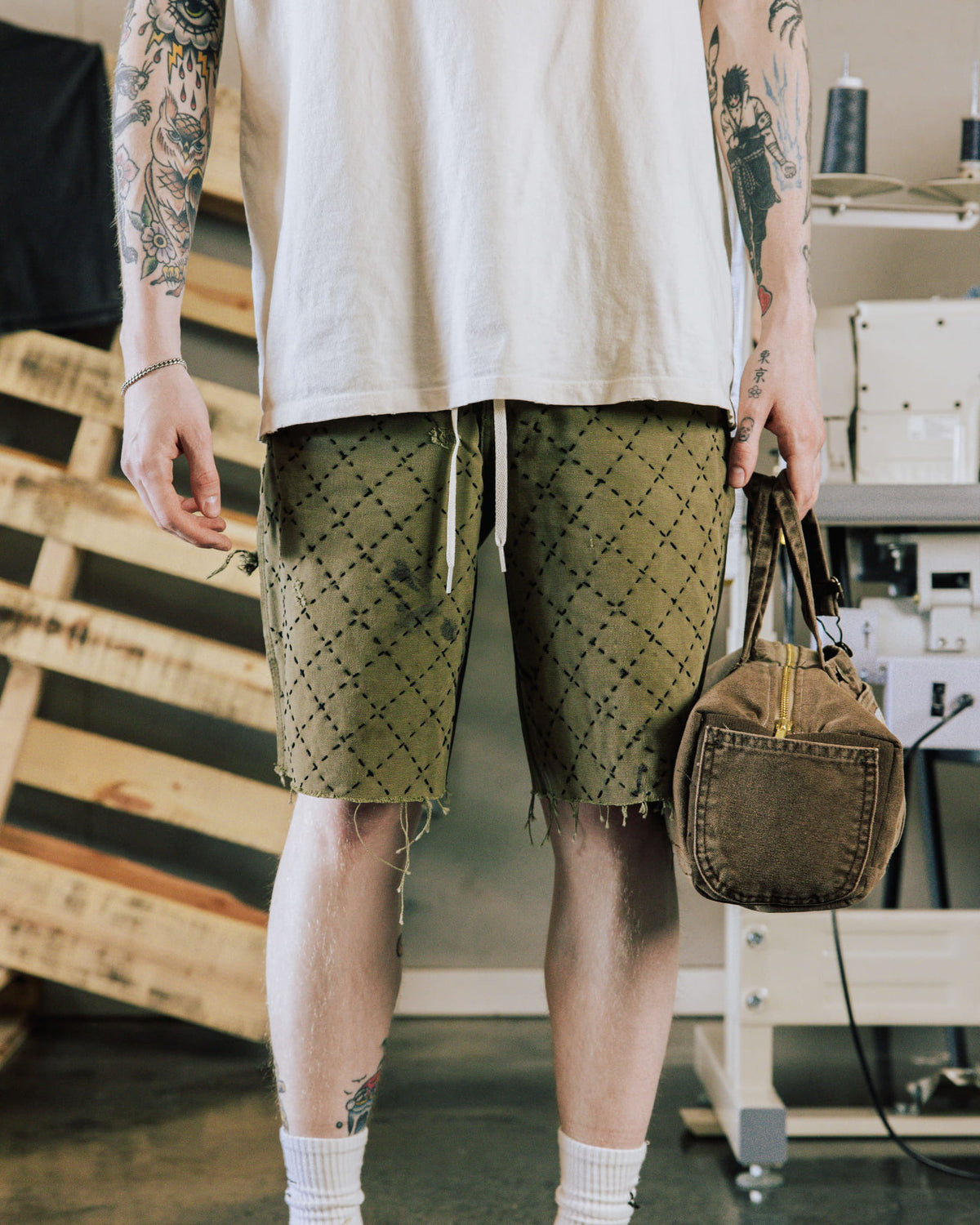 Tattooed person wears olive patterned drawstring shorts with frayed hems and a white T-shirt, holding a brown canvas bag.