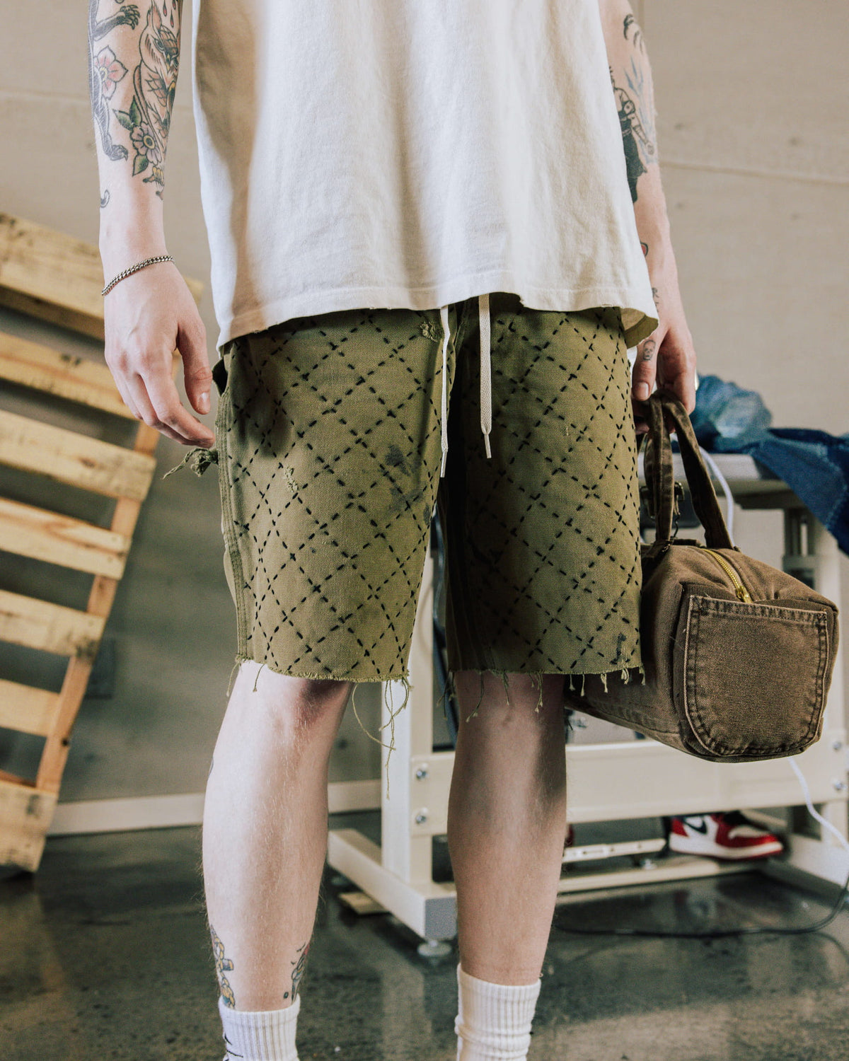 Person wearing olive drawstring shorts with black diamond stitching and frayed hems, a white tee, and holding a brown bag.